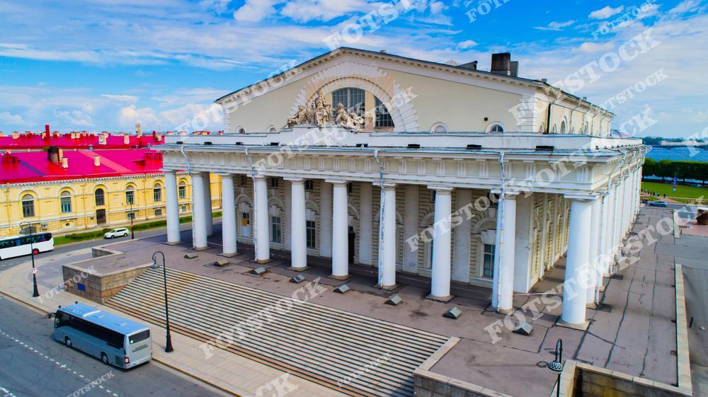 На фотографии изображено здание Биржи на Стрелке Васильевского острова в Санкт-Петербурге, построенное в стиле классицизма. Оно является одним из символов города. Массивные колонны и симметричная композиция здания придают ему величественный вид. В летний день фасад здания освещен ярким солнечным светом, что подчеркивает его архитектурные детали.